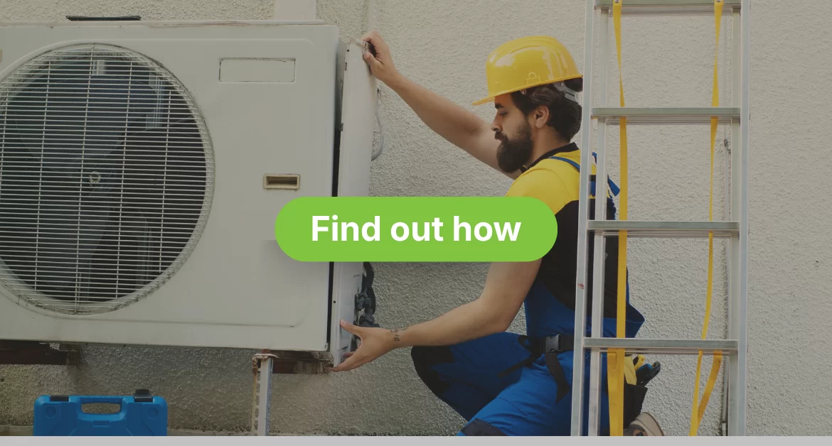 A maintenance technician with a beard is kneeling beside an outdoor air conditioner unit, working on disassembling or fixing it. He is wearing a blue uniform, a yellow and black shirt, and a yellow hard hat. A silver ladder is visible on the right, and a blue toolbox is on the ground to the left. The AC unit is mounted on a white, textured wall.