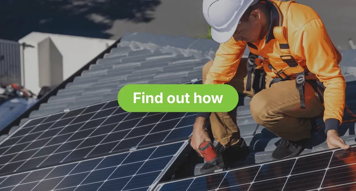 A solar panel installer, wearing a white hard hat, an orange high-visibility long-sleeved shirt, brown pants, and a safety harness, is crouched on a residential roof. The technician is using a power tool to secure or work on a large array of dark solar panels. The background shows surrounding suburban homes and rooftops under a bright sky.