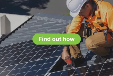 A solar panel installer, wearing a white hard hat, an orange high-visibility long-sleeved shirt, brown pants, and a safety harness, is crouched on a residential roof. The technician is using a power tool to secure or work on a large array of dark solar panels. The background shows surrounding suburban homes and rooftops under a bright sky.