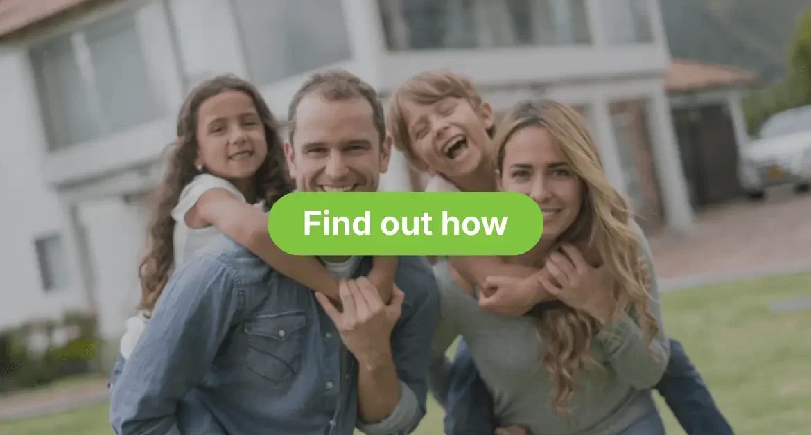 A smiling family of four standing outside on a lawn in front of a modern, blurred house. The father, wearing a denim shirt, is giving the young daughter a piggyback ride. The mother, wearing a grey top, is giving the young son a piggyback ride; the son is laughing enthusiastically.