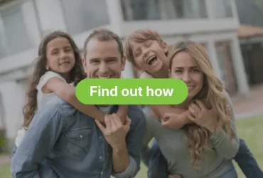 A smiling family of four standing outside on a lawn in front of a modern, blurred house. The father, wearing a denim shirt, is giving the young daughter a piggyback ride. The mother, wearing a grey top, is giving the young son a piggyback ride; the son is laughing enthusiastically.
